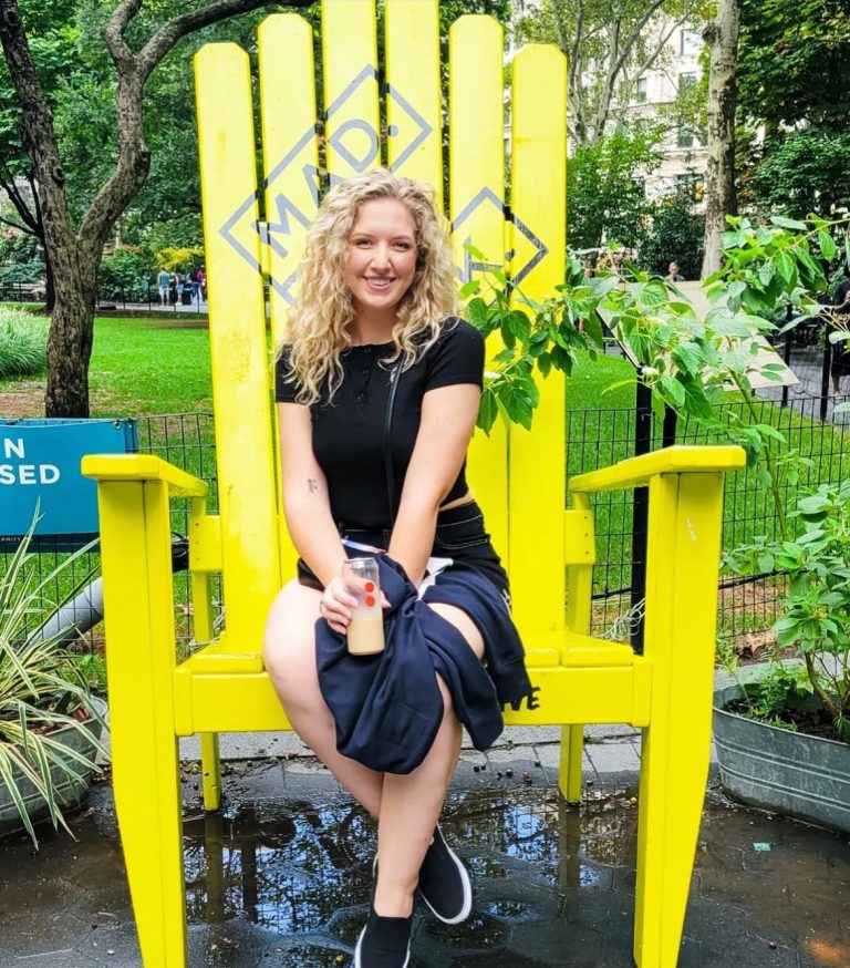 Woman seated in an oversized bright yellow XXL wooden chair in a green city park