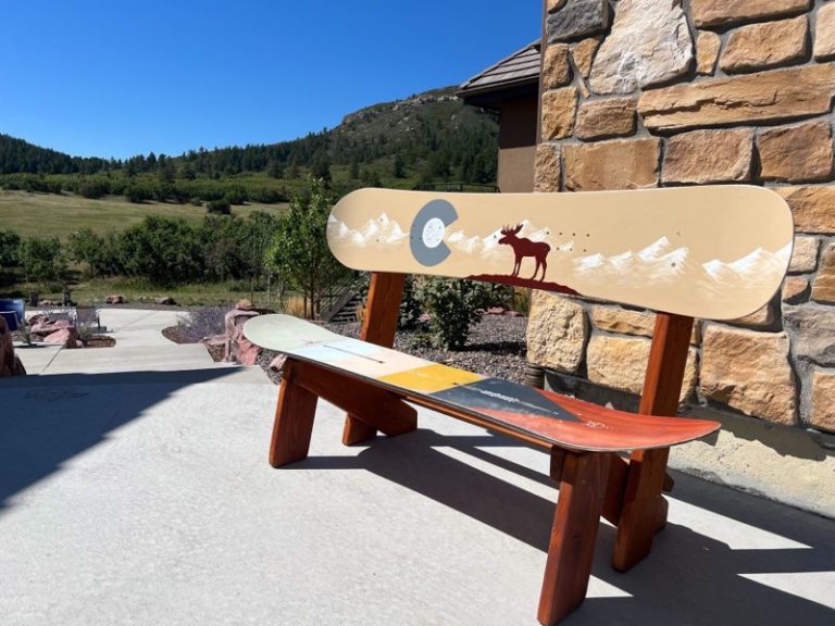 Snowboard bench with a painted moose, white peaks and Colorado flag C, beside a stone wall