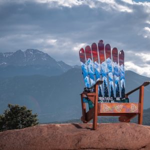 Ski chair painted with snowy peaks and pine forest, perched on red rock above the Rockies