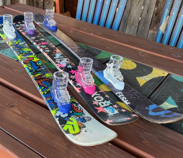 Shot skis with clear ski-boot shot glasses mounted on colorful graphic skis, laid across a wood deck