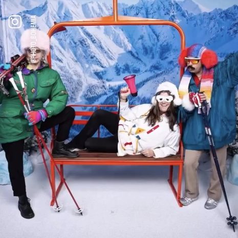 Three-person orange replica chairlift bench anchoring a snowy photo booth set with costumed skiers