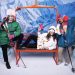 Three-person orange replica chairlift bench anchoring a snowy photo booth set with costumed skiers