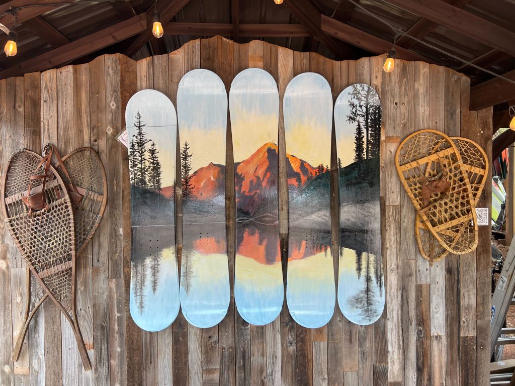 Five snowboard decks forming a sunset mountain lake mural on barnwood, flanked by antique snowshoes