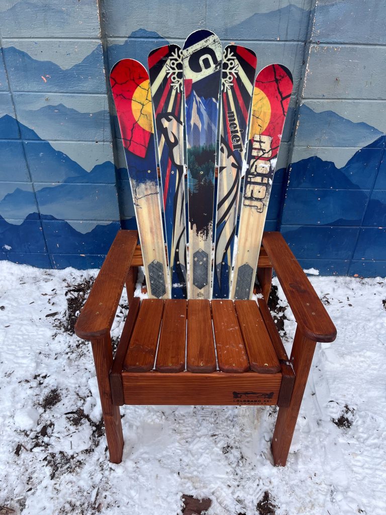 Tall compact alpine chair backed with colorful Colorado flag skis, standing in fresh snow