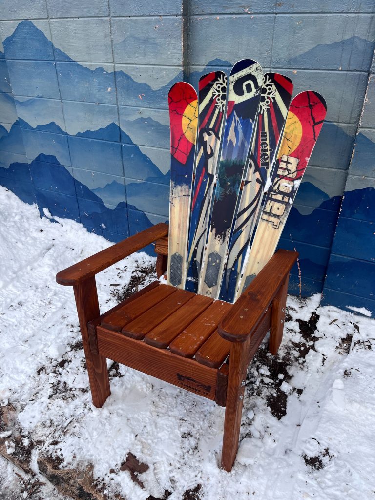 Alpine ski chair built from skis, standing in the snow in front of a hand painted mountain mural